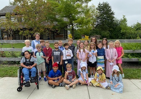 Elementary students pose on field trip in front of log cabin